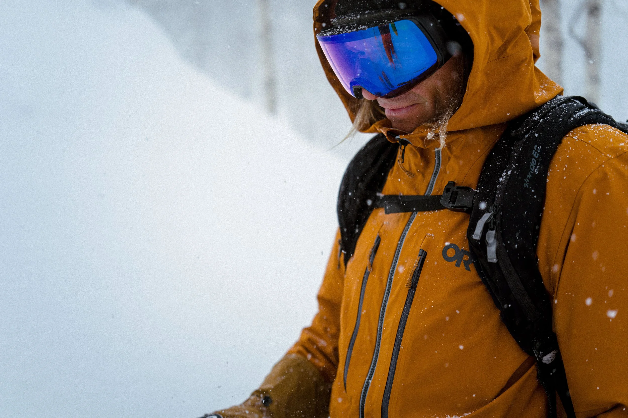 A person in a yellow winter jacket with a black backpack, wearing a blue reflective ski or snowboarding goggles, is in a snowy outdoor environment.