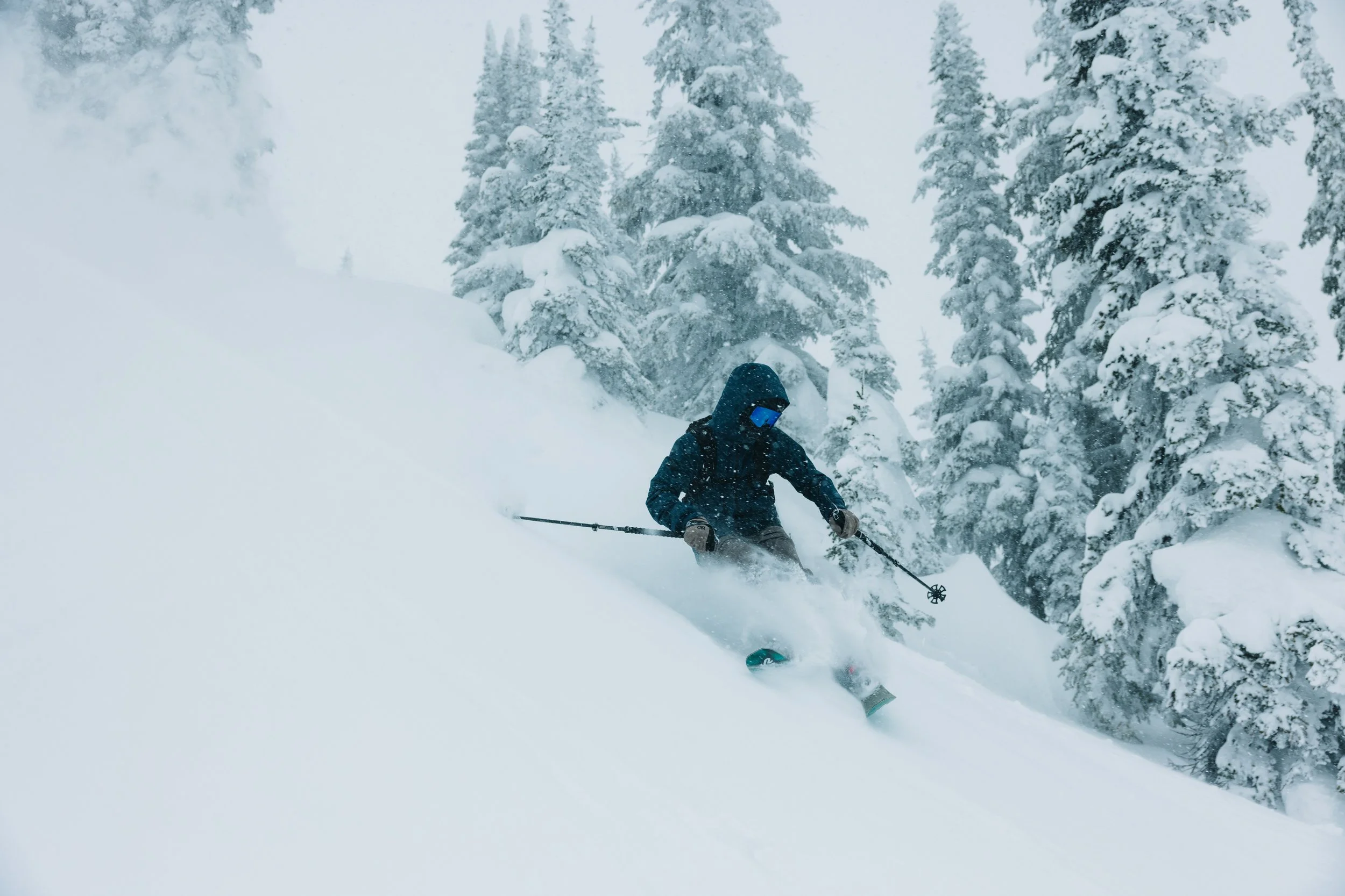 A person skiing on a snow-covered slope surrounded by snow-laden pine trees during a snowy day.