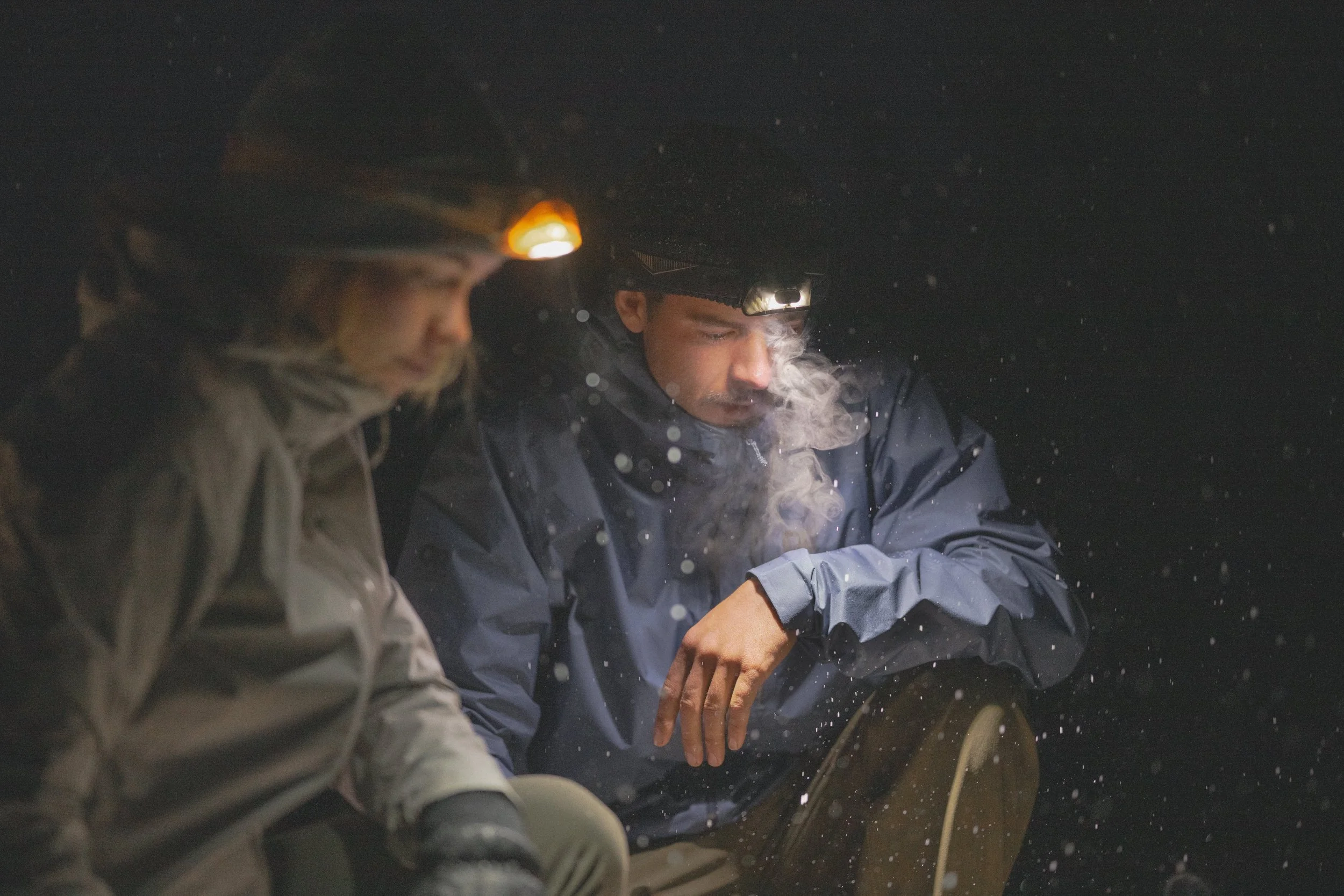 Two people wearing headlamps and outdoor jackets in the dark, with visible breath and snow or frost in the air.