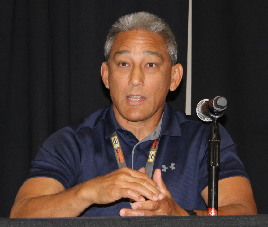 A middle-aged man with short gray hair speaking at a panel, sitting behind a table with a microphone, wearing a navy blue polo shirt and a lanyard.