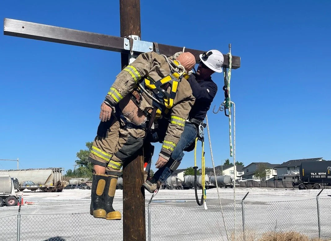 A firefighter in full gear and a construction worker in a hard hat and safety harness performing a rescue on a utility pole against a clear blue sky.