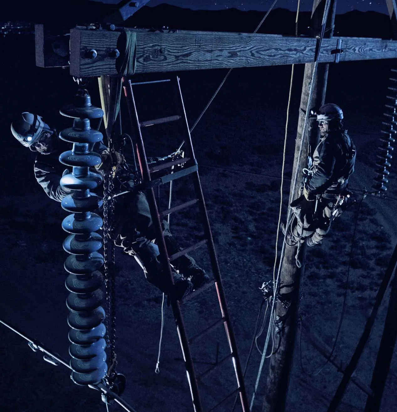 Two workers wearing safety harnesses and helmets are working on electrical lines at night, climbing a wooden utility pole with various equipment attached.
