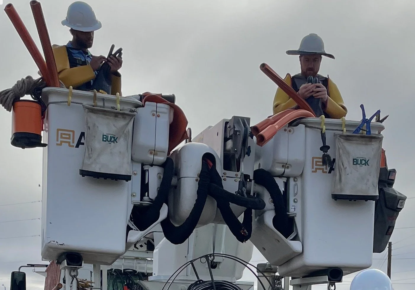 Two utility workers in buckets on a bucket truck, wearing safety helmets and yellow jackets, working on electrical lines.