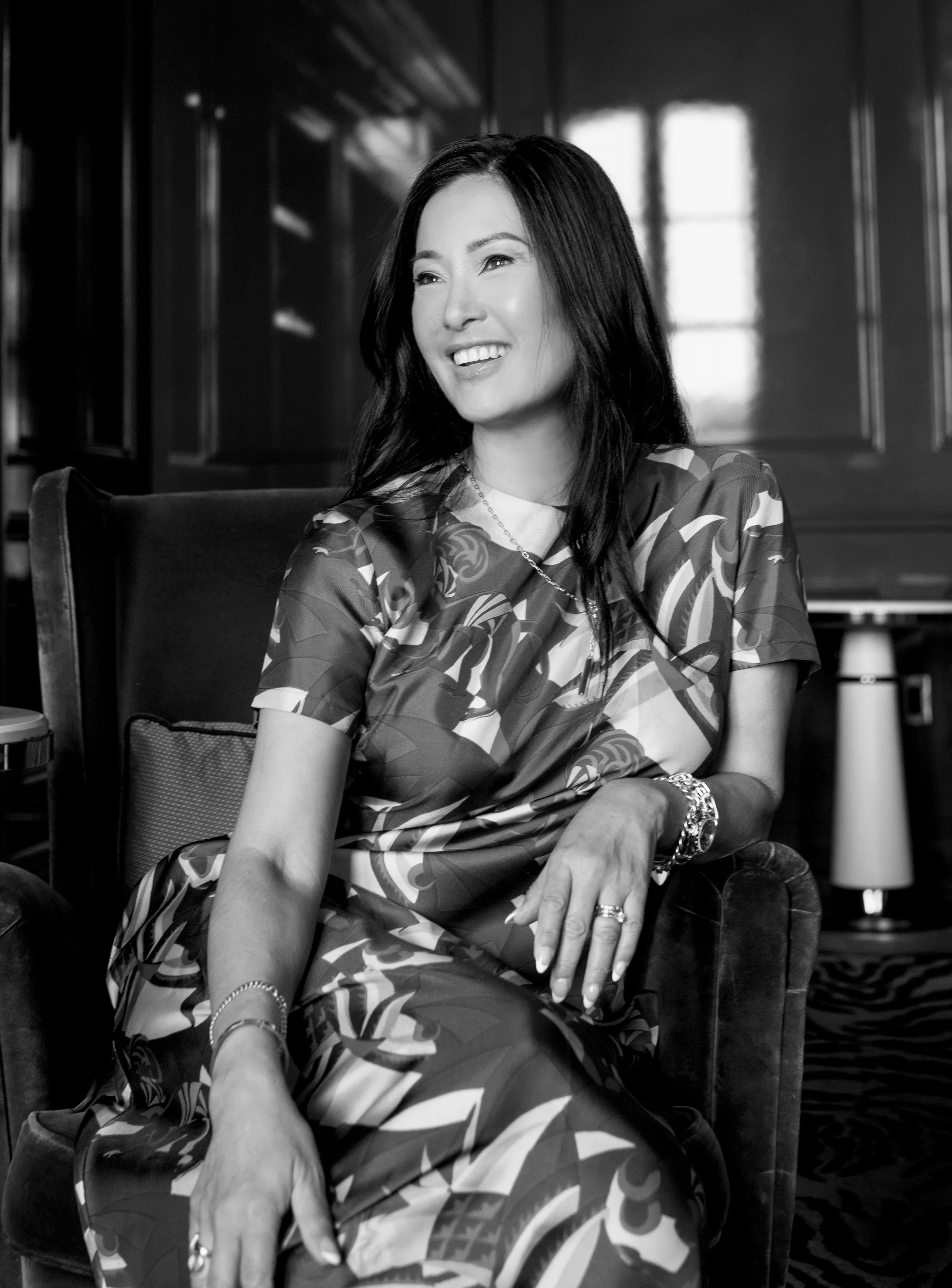 A woman sitting on a chair in a room with wooden paneled walls and a window, smiling and looking to her left, in black and white.