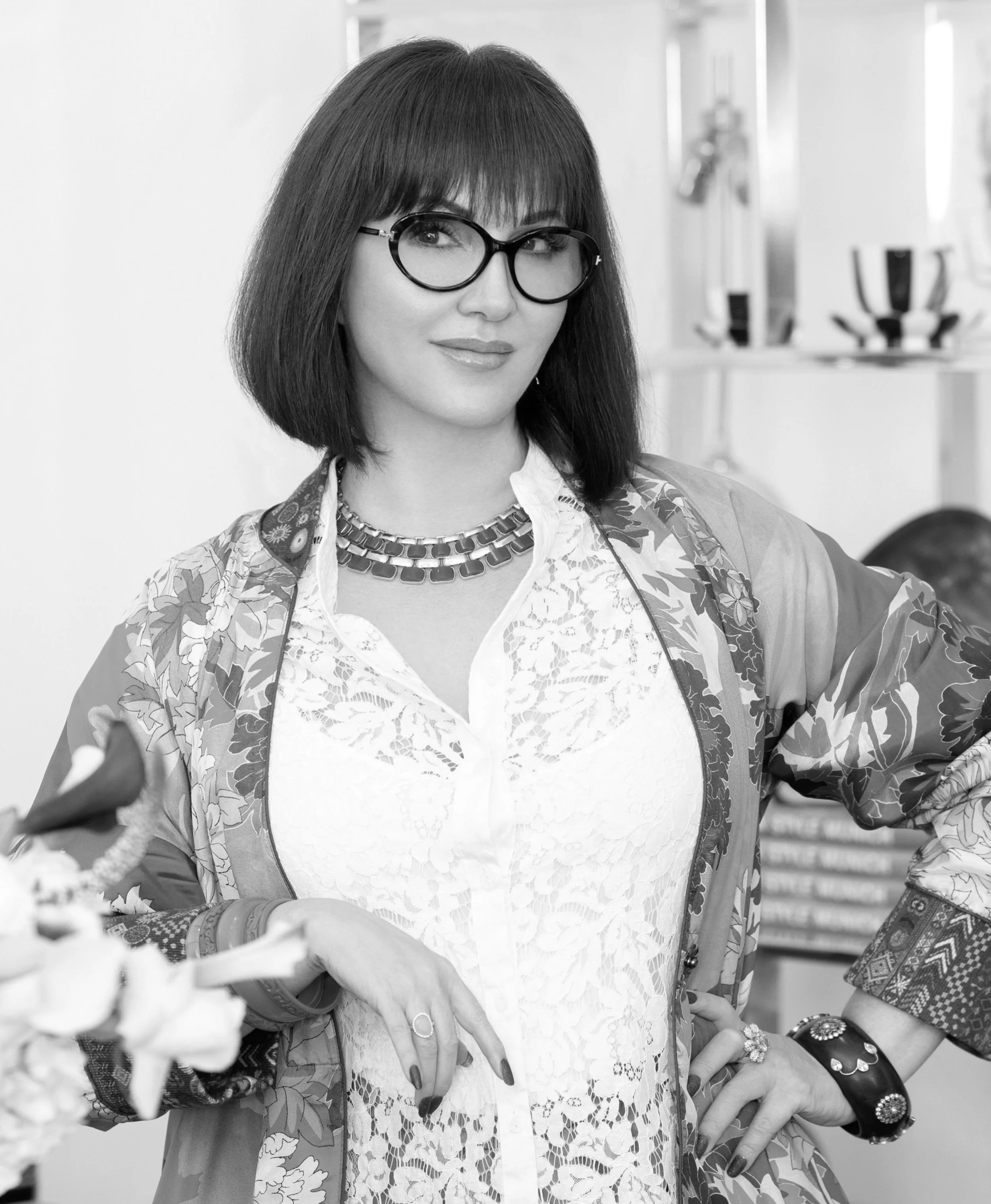 A woman with shoulder-length dark hair and glasses, wearing a patterned jacket over a white lace blouse, standing indoors with her hands on her hips.