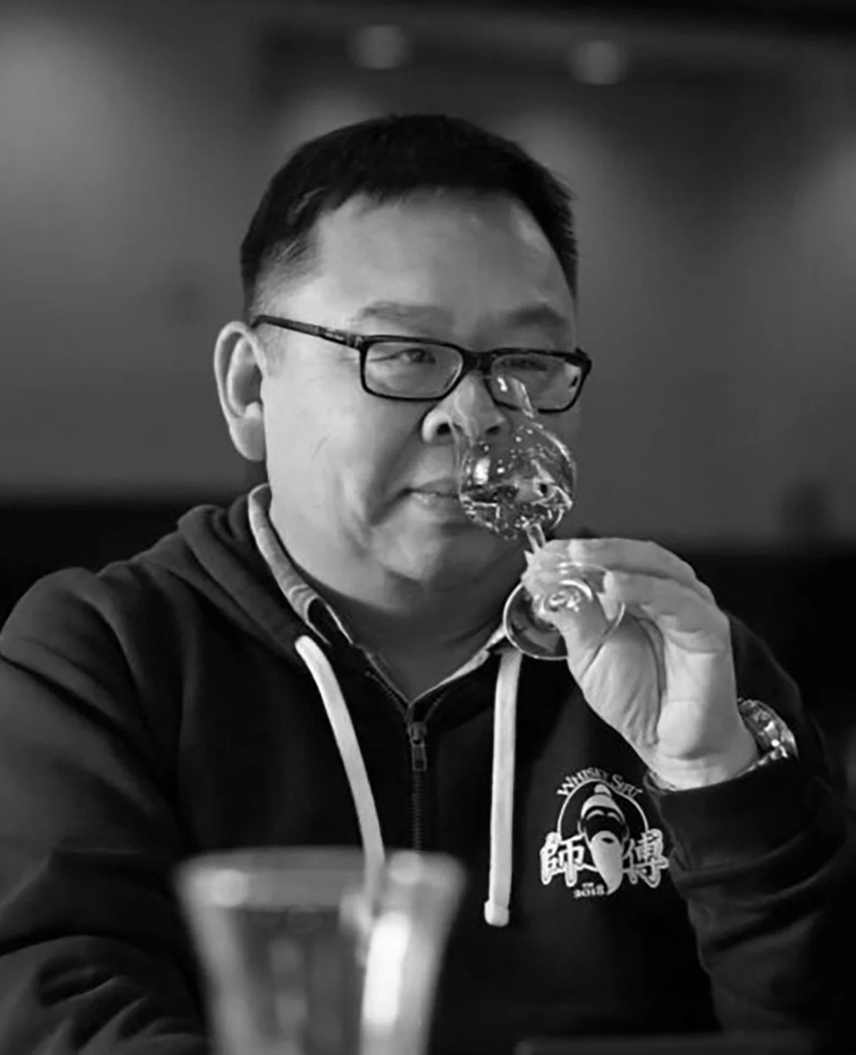 Man wearing glasses and a hoodie, holding a wine glass, tasting a beverage in a restaurant or bar setting.