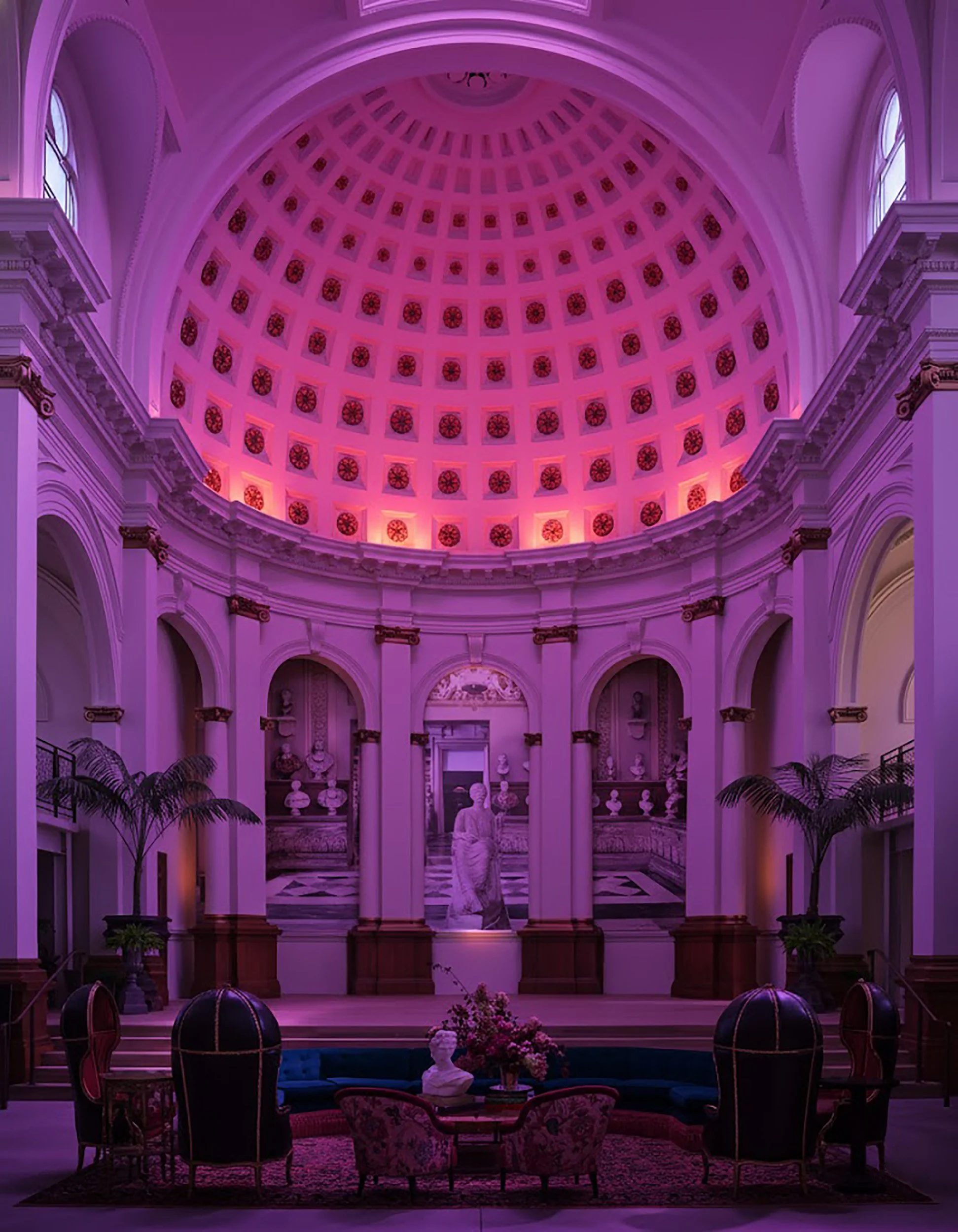 Interior of a grand hall with a high, domed ceiling illuminated in purple, featuring classical architecture and statue decor.