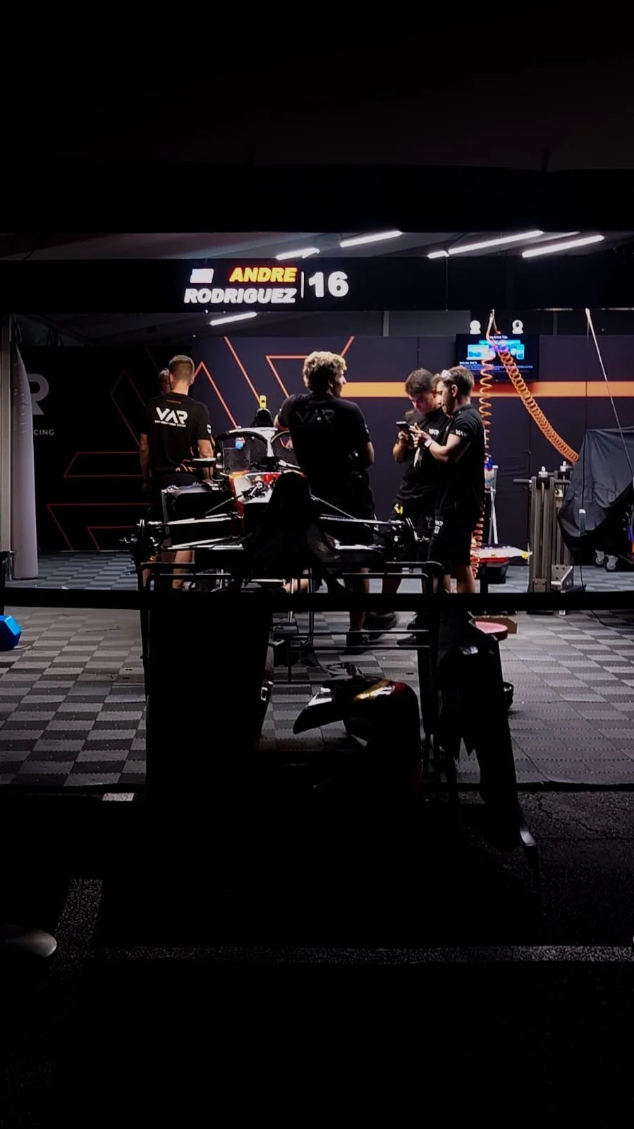 Race car garage with mechanics and team members gathered around a vehicle, illuminated by bright artificial lighting, with a sign showing the name 'Andre Rodriguez 16.'