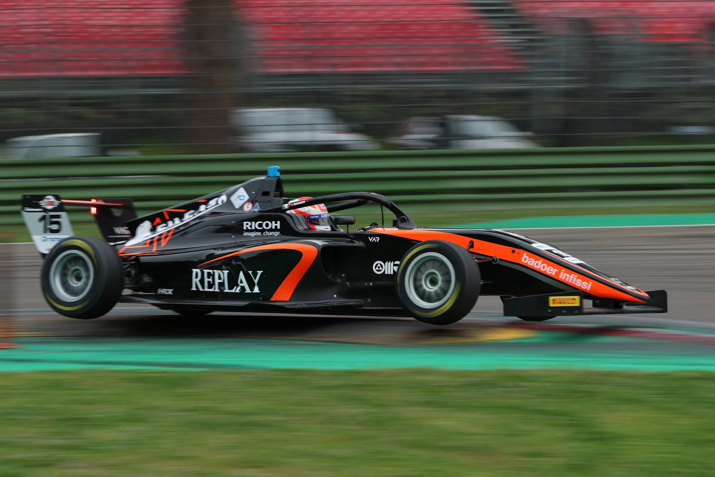 A black and orange Formula 2 race car speeding on a race track, with sponsor logos including Replay, Ricoh, and Pirelli, and the number 15 on the rear wing.