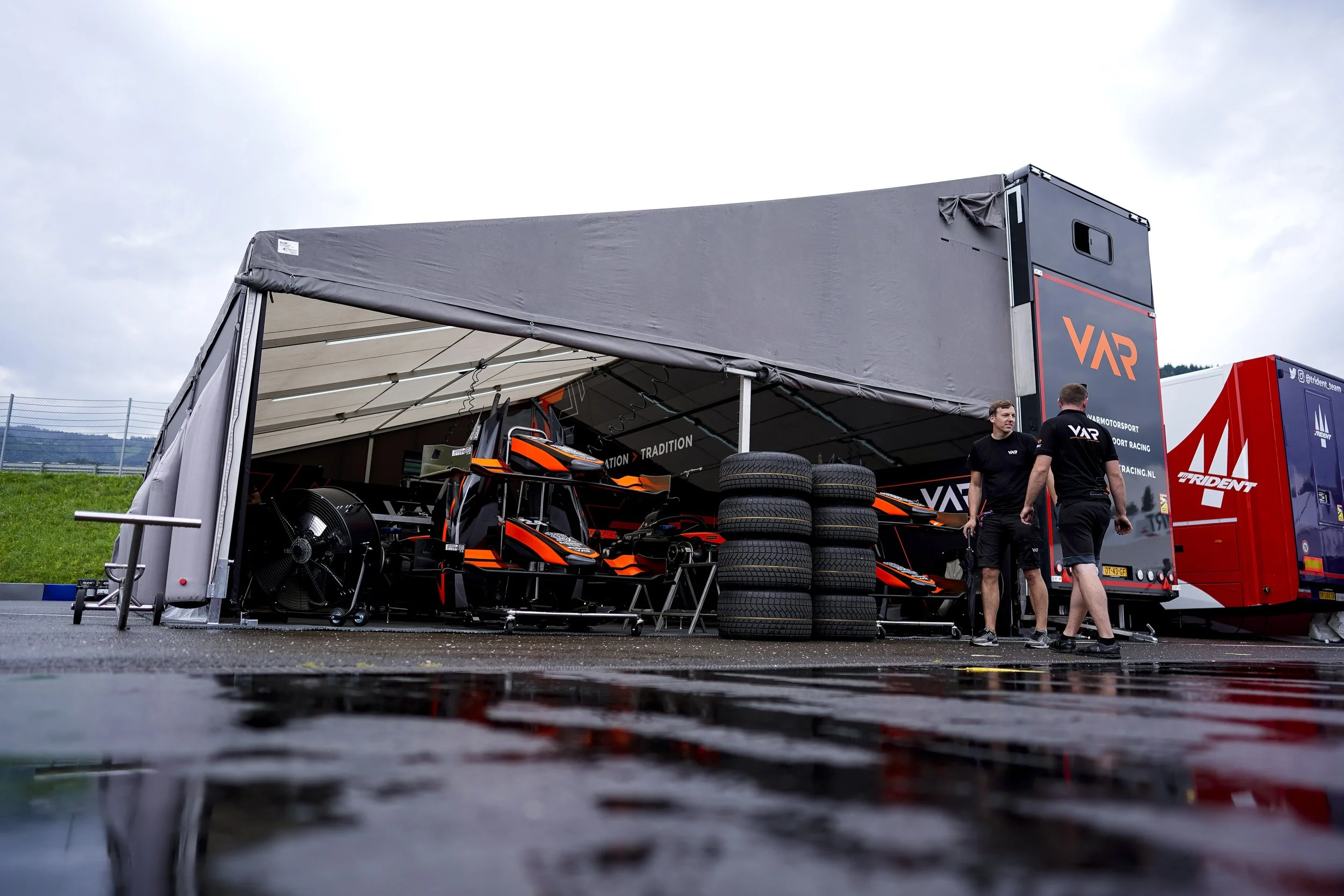 Race car garage with two orange and black single-seater race cars, tires stacked next to them, and two crew members in black shirts talking, set up on wet track with a trailer in the background.