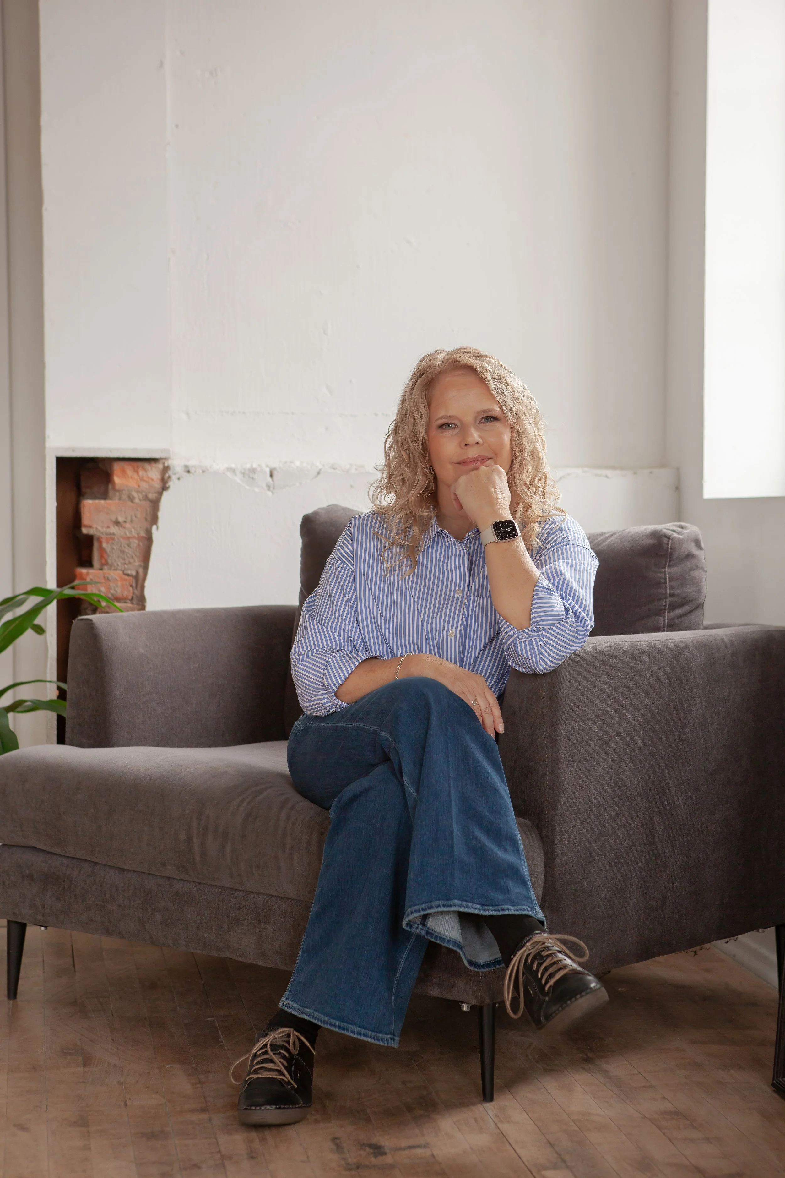 A woman with curly blonde hair sitting on a gray couch in a room with white walls, wearing a blue and white striped shirt, blue jeans, and black shoes. She has a watch on her left wrist and is resting her chin on her right hand, looking at the camera.