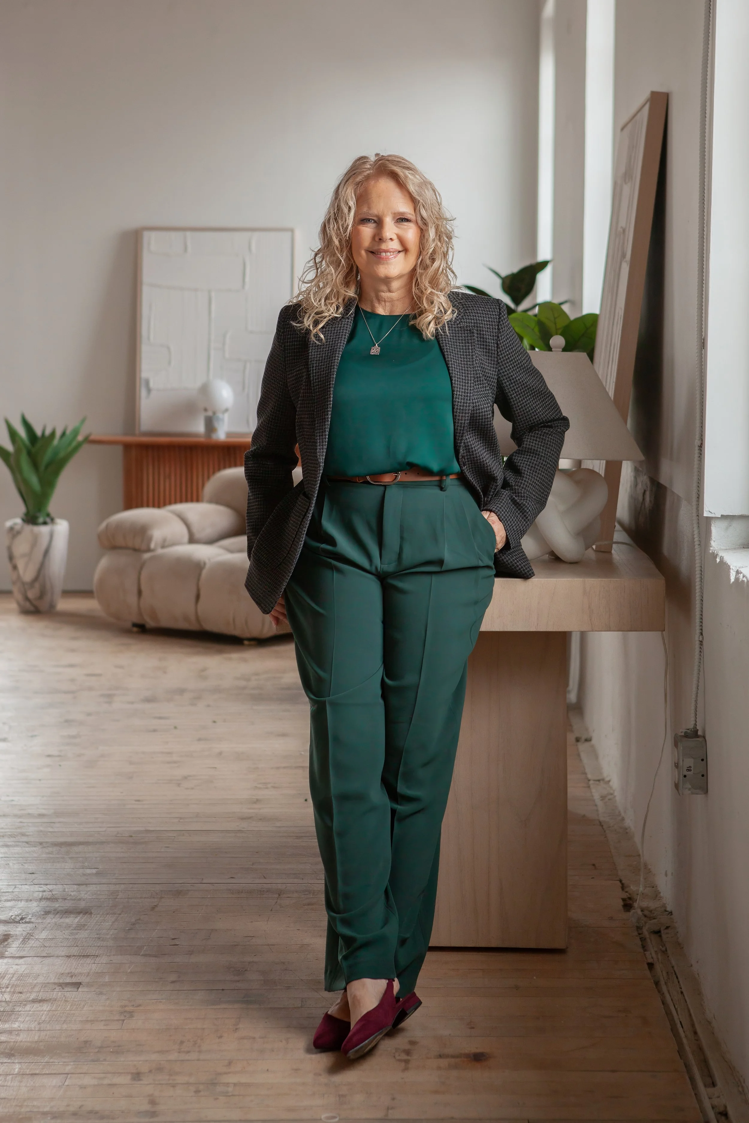 A smiling woman with curly blonde hair standing in a modern, well-lit interior, leaning against a wooden table with her hands in her pockets, wearing a dark blazer, teal blouse, green pants, and burgundy shoes.