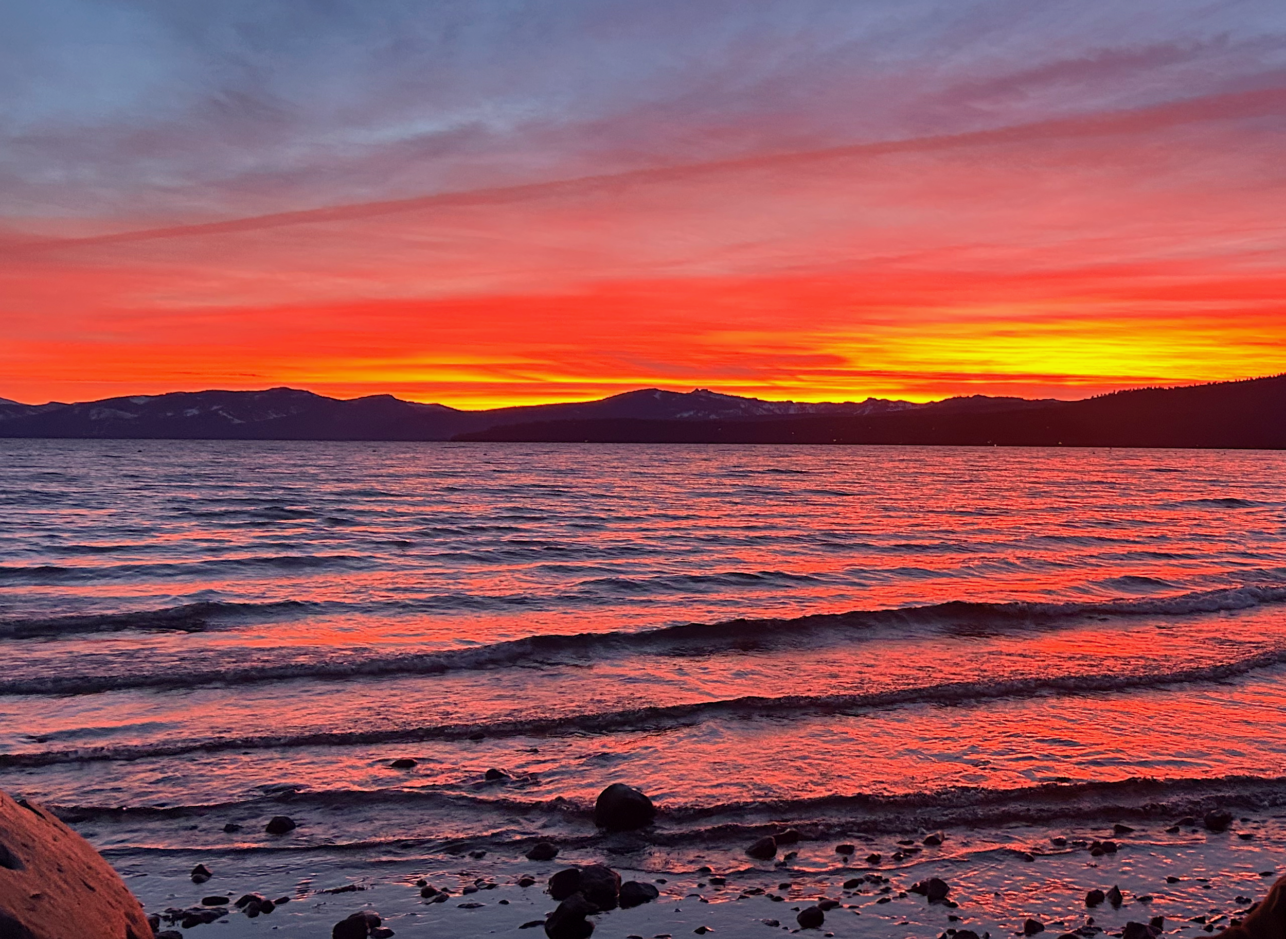 Colorful sunset over a lake with mountains in the background, reflecting pink and orange hues on the water and sky.