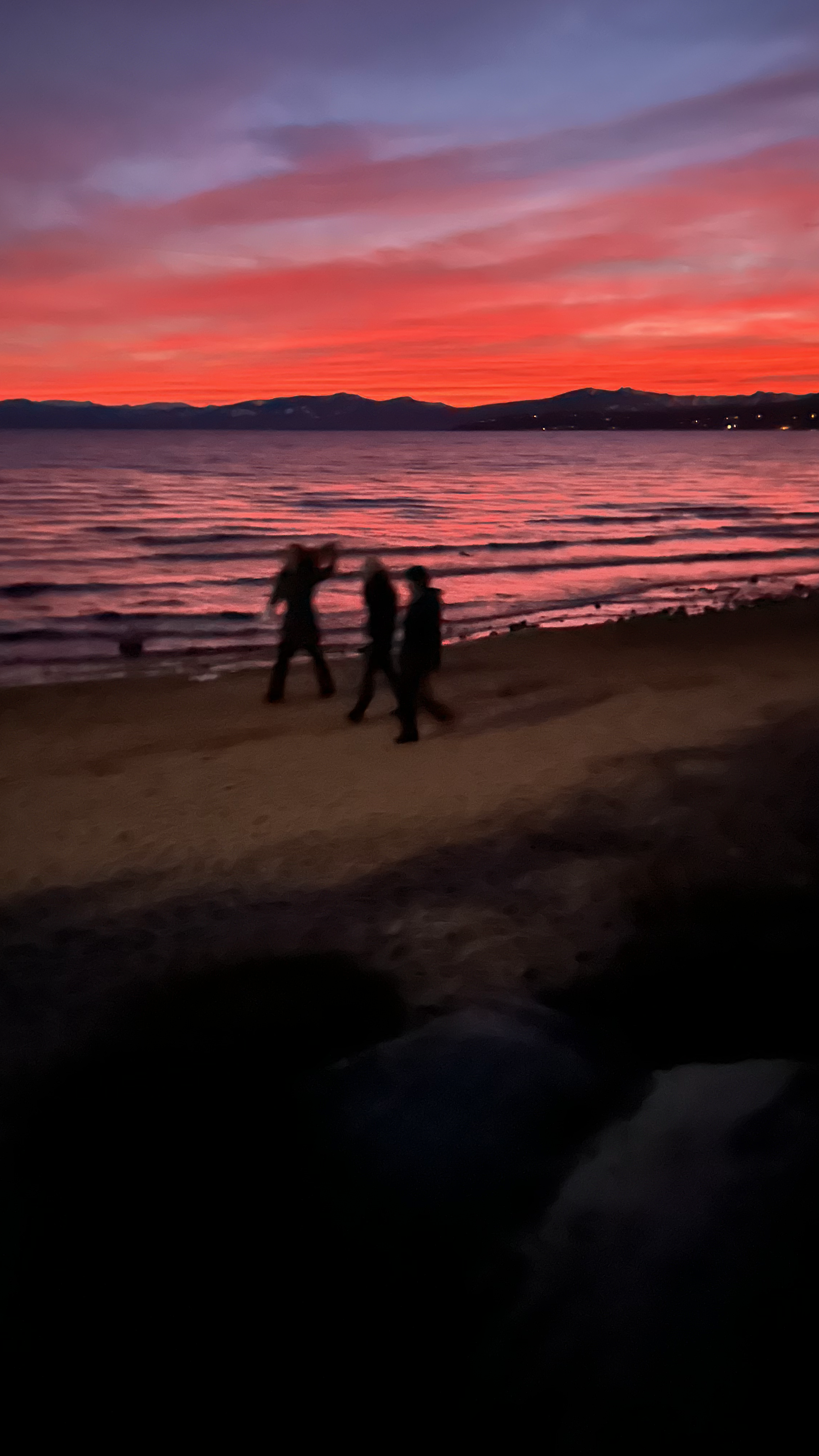 Silhouettes of three people walking on a beach during sunset, with colorful pink and purple sky and mountains in the background.