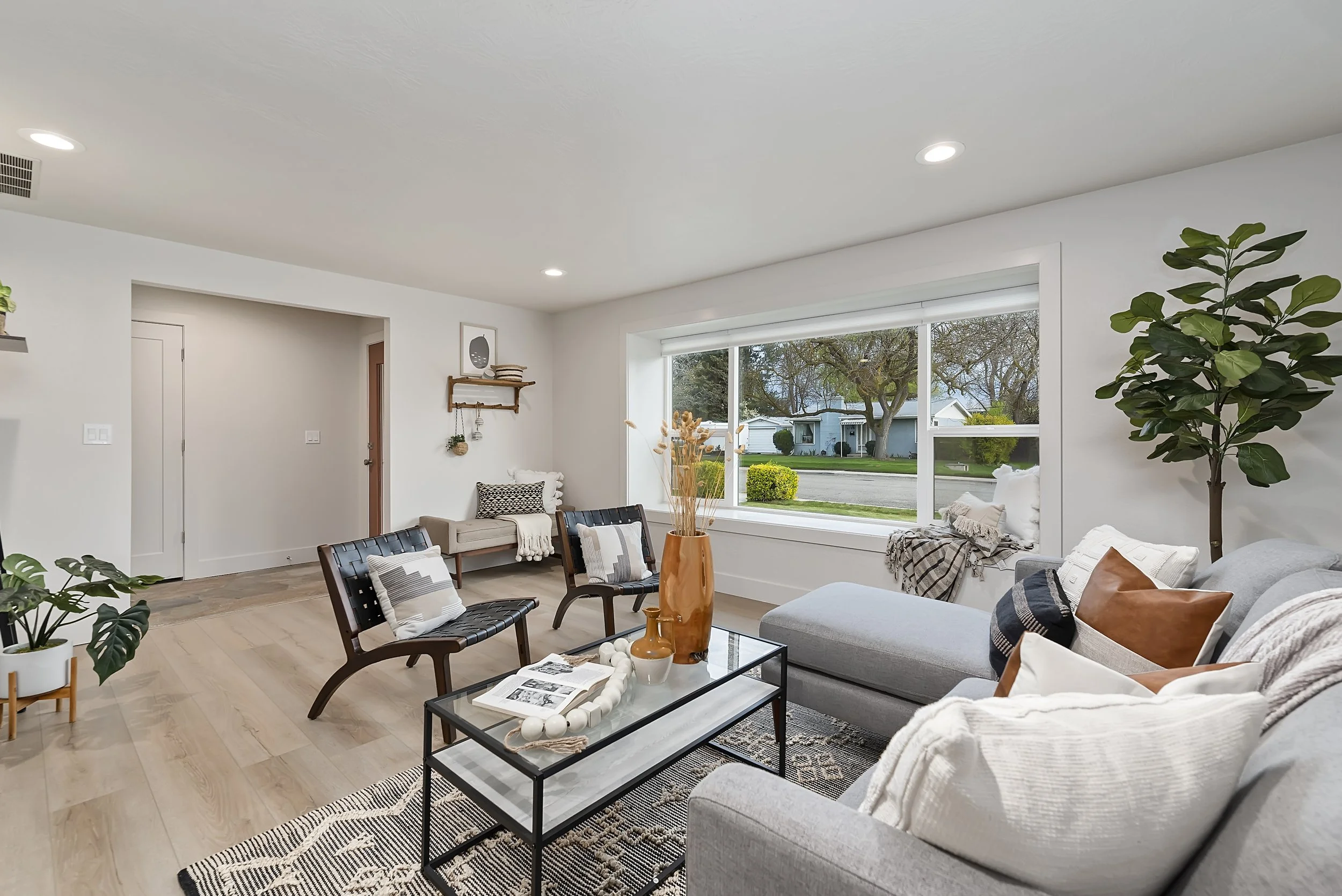 Bright, modern living room with large window, gray sectional sofa, black chairs, wooden coffee table, and decorative plants.