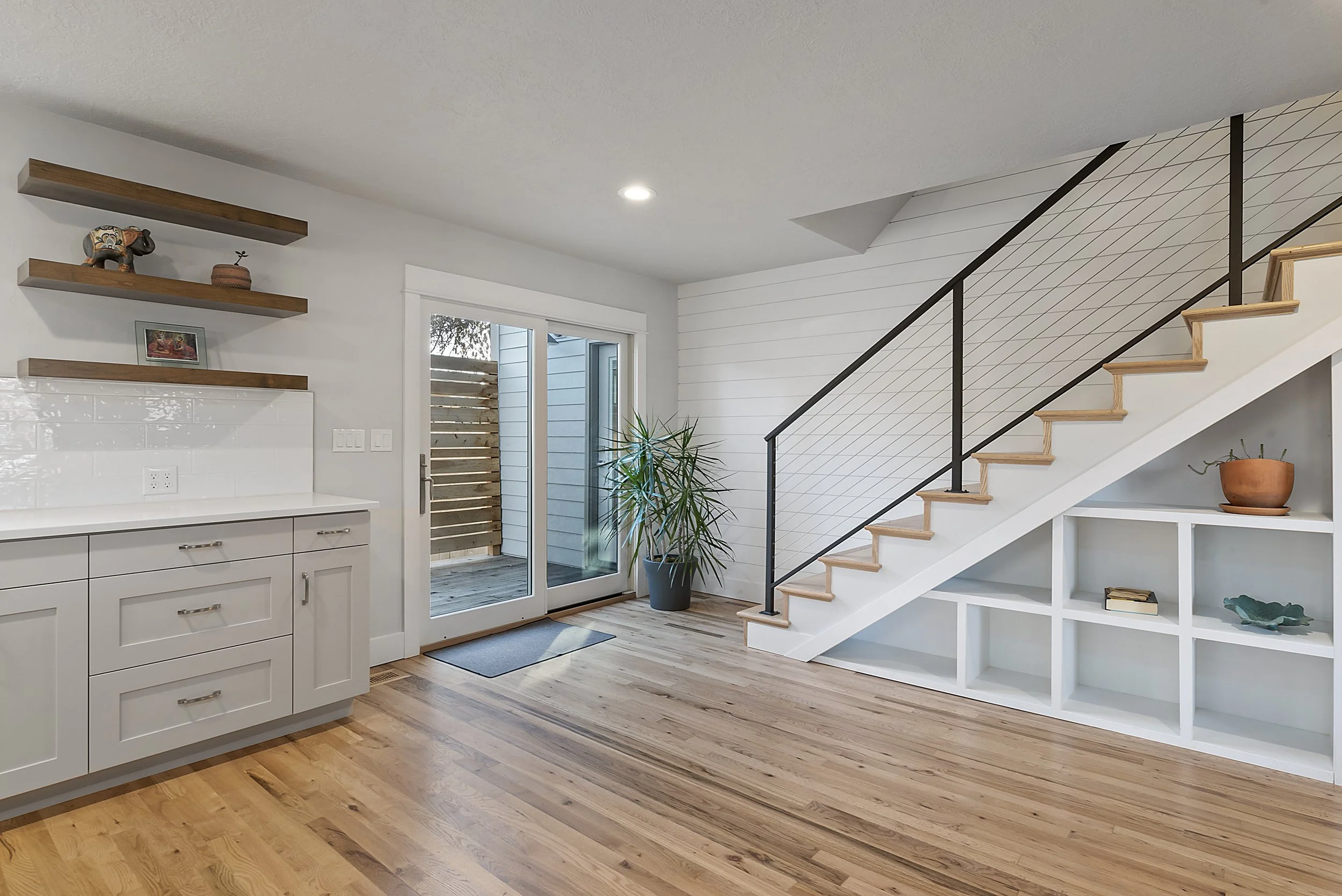 Modern living space with white cabinets, open shelves, a sliding glass door, wooden floor, potted plant, and a staircase with built-in shelving underneath.