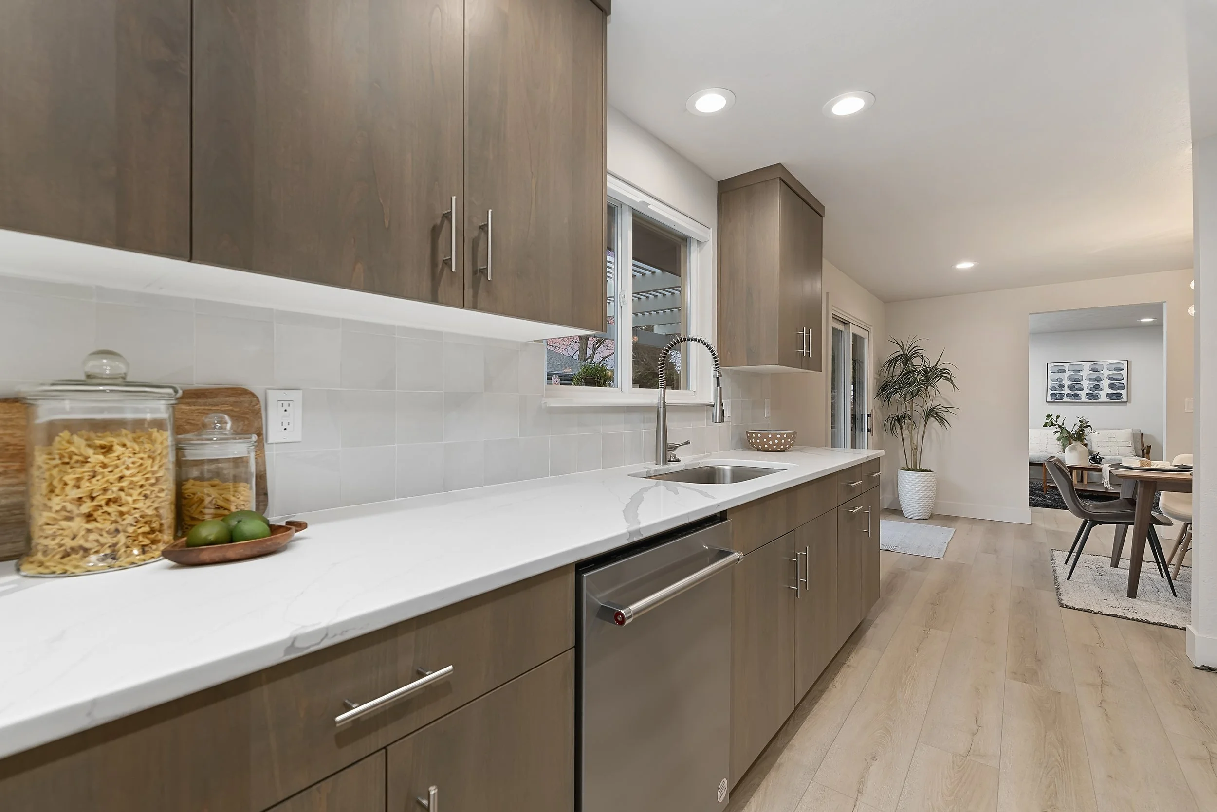 Modern kitchen with light wood cabinets, white countertop, tiled backsplash, stainless steel dishwasher, and a window above the sink. The space opens into a dining area with a table and chairs, and a living room in the background.