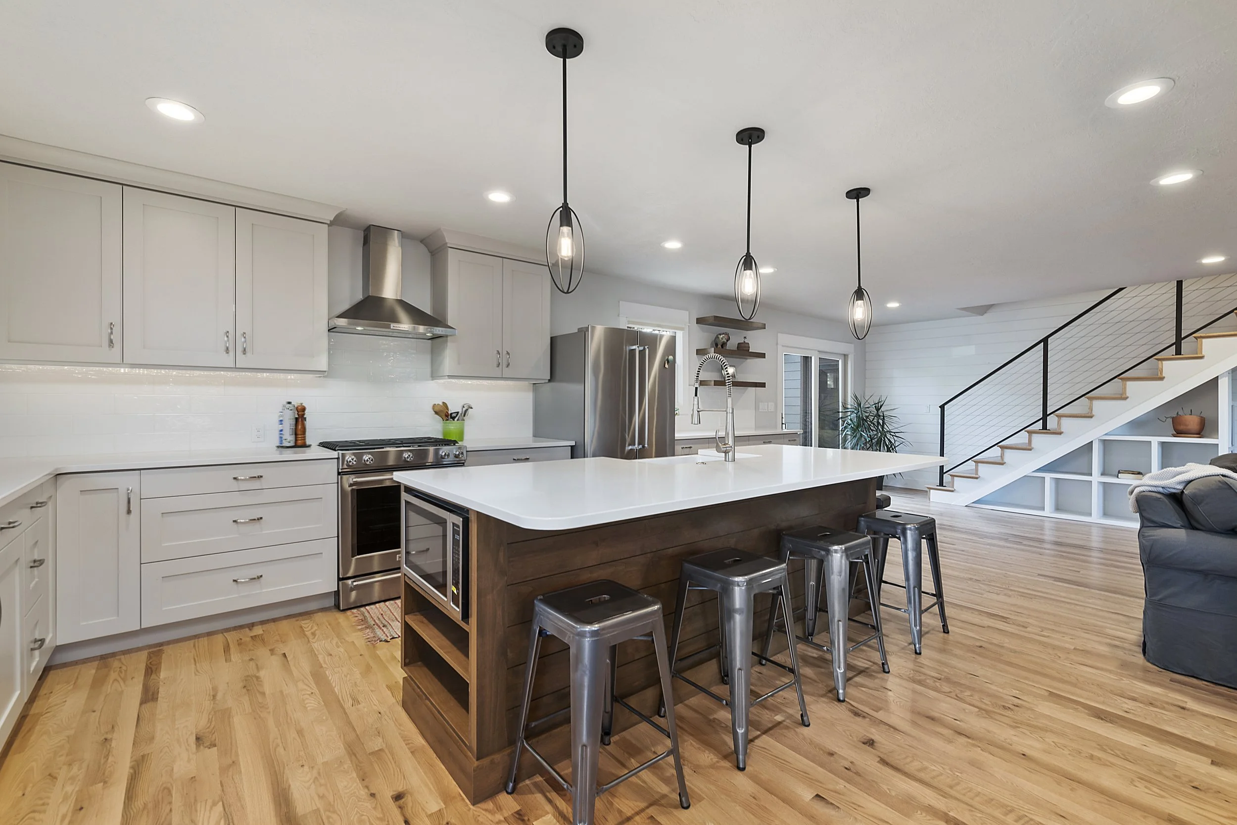 Modern kitchen with white cabinets, stainless steel appliances, a large white island with a wooden base, and four metal barstools. There are three pendant lights hanging above the island, hardwood floors, and an open living space with stairs leading to an upper level.