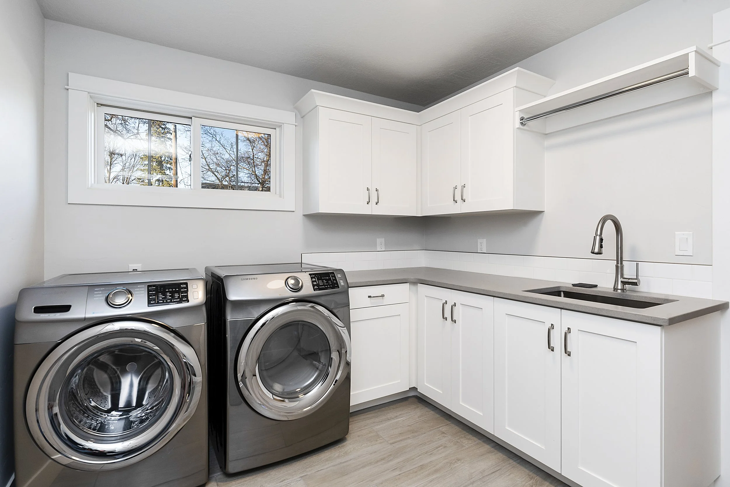 Modern laundry room with white cabinets, grey countertops, a stainless steel sink, a large window showing trees outside, and a front-loading washer and dryer.