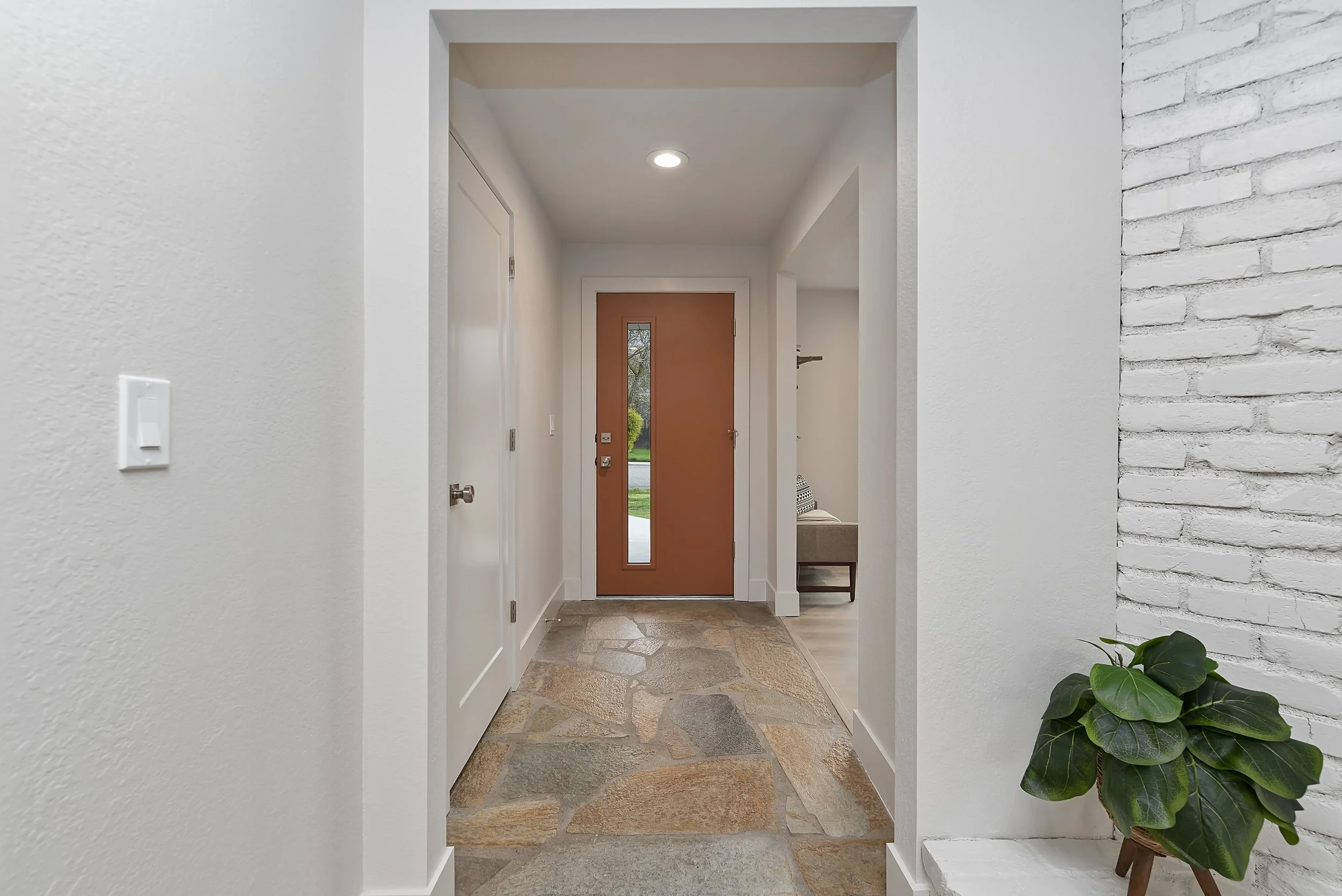 Interior view of a modern home entryway with a wooden front door, stone flooring, white walls, and a potted plant on a small ledge.