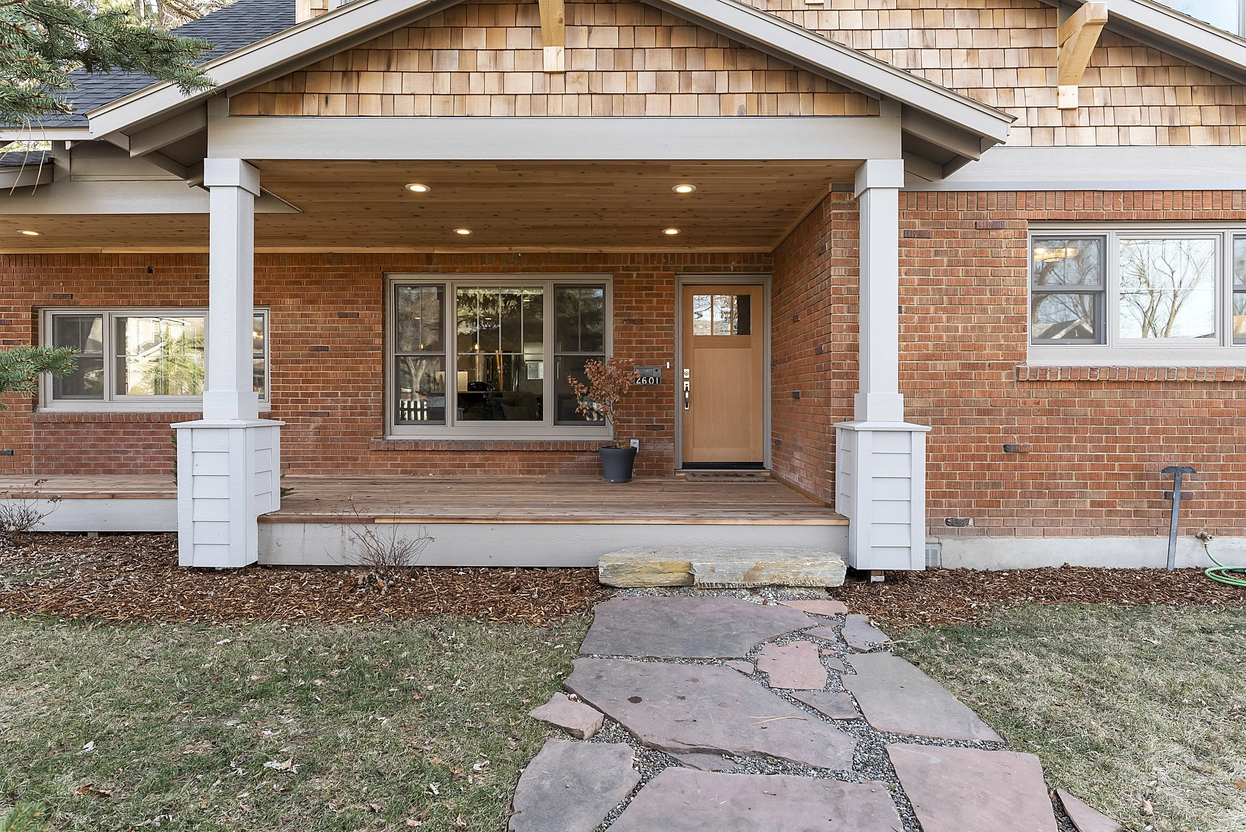 Front porch of a brick house with wooden flooring, white pillars, and a wooden front door. There is a potted plant next to the door and a stone pathway leading to the stairs.