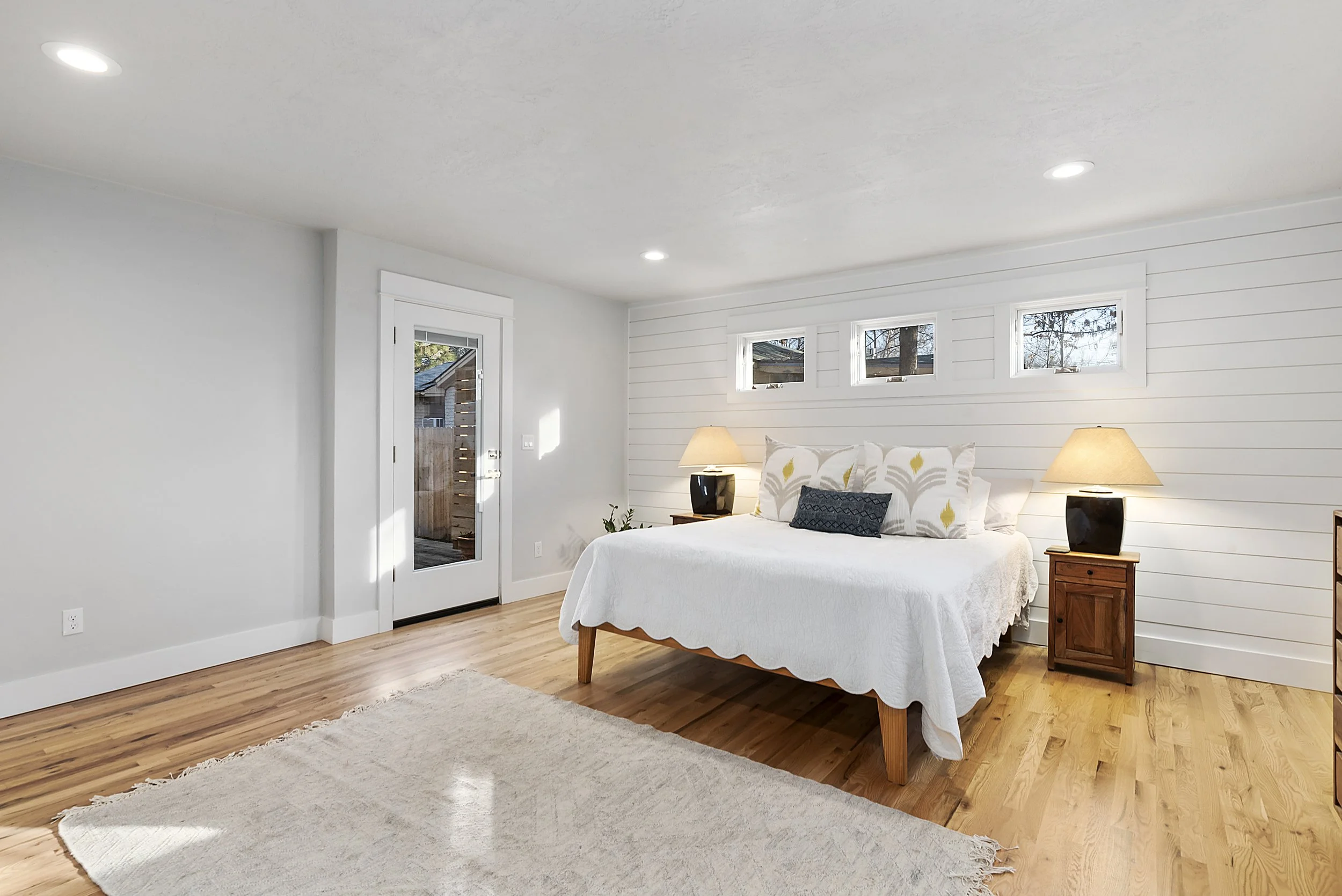 A bedroom with a white bedspread, two bedside tables with lamps, a light wood floor, and a white wall with small horizontal windows.