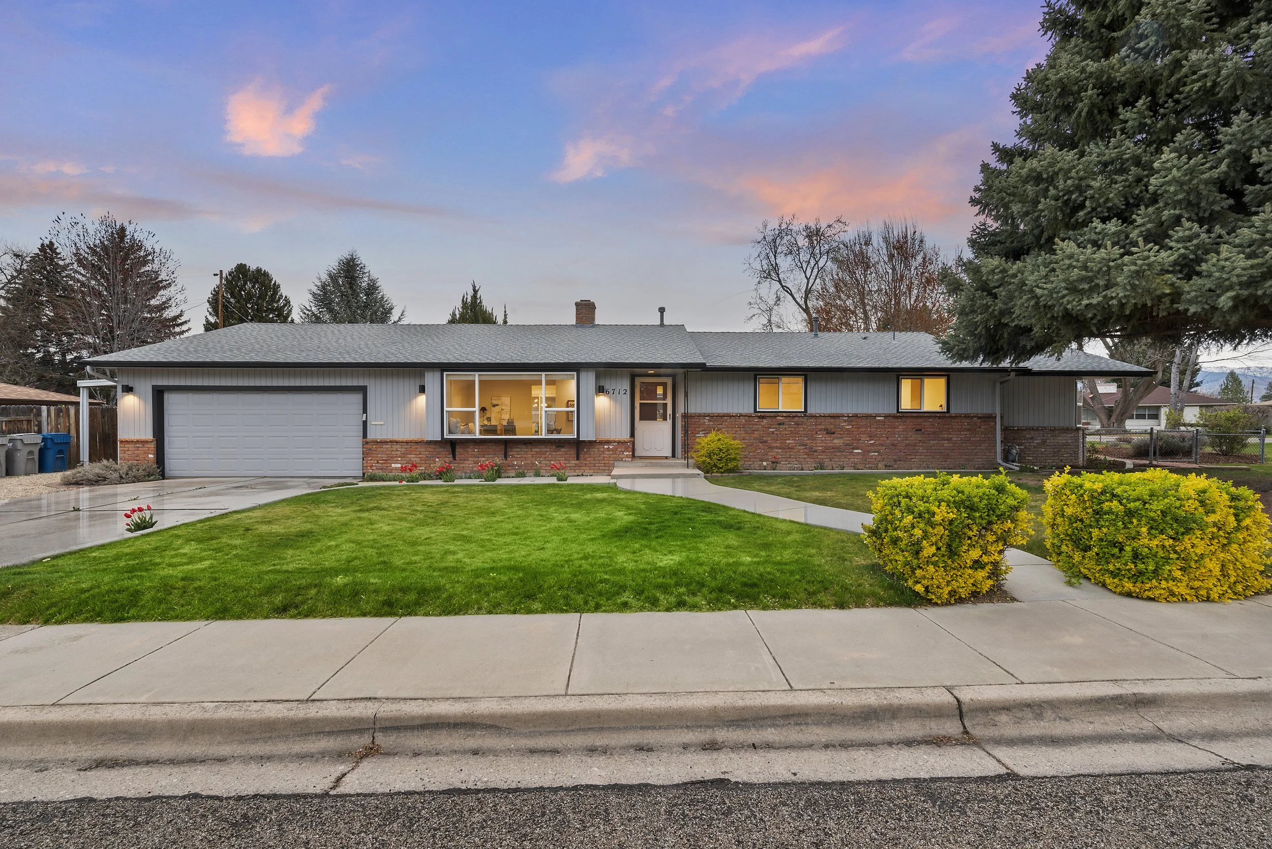 Front view of a single-story house with a gray exterior, brick accents, and a attached garage. The house is surrounded by a well-maintained lawn, bushes, and trees, with pink flowers along the sidewalk. The sky is showing a pink and blue sunset.