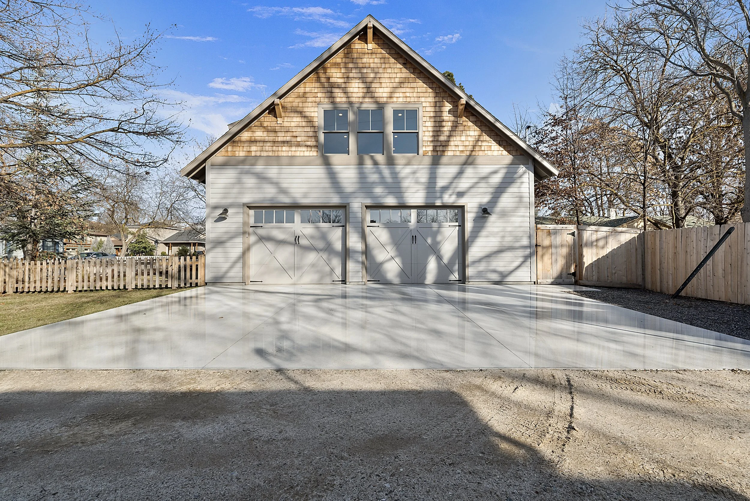 A newly built two-car garage with white doors and a light gray exterior, situated in a residential neighborhood with a concrete driveway, wooden fencing on the sides, and trees with bare branches, in a sunny day with a clear blue sky.