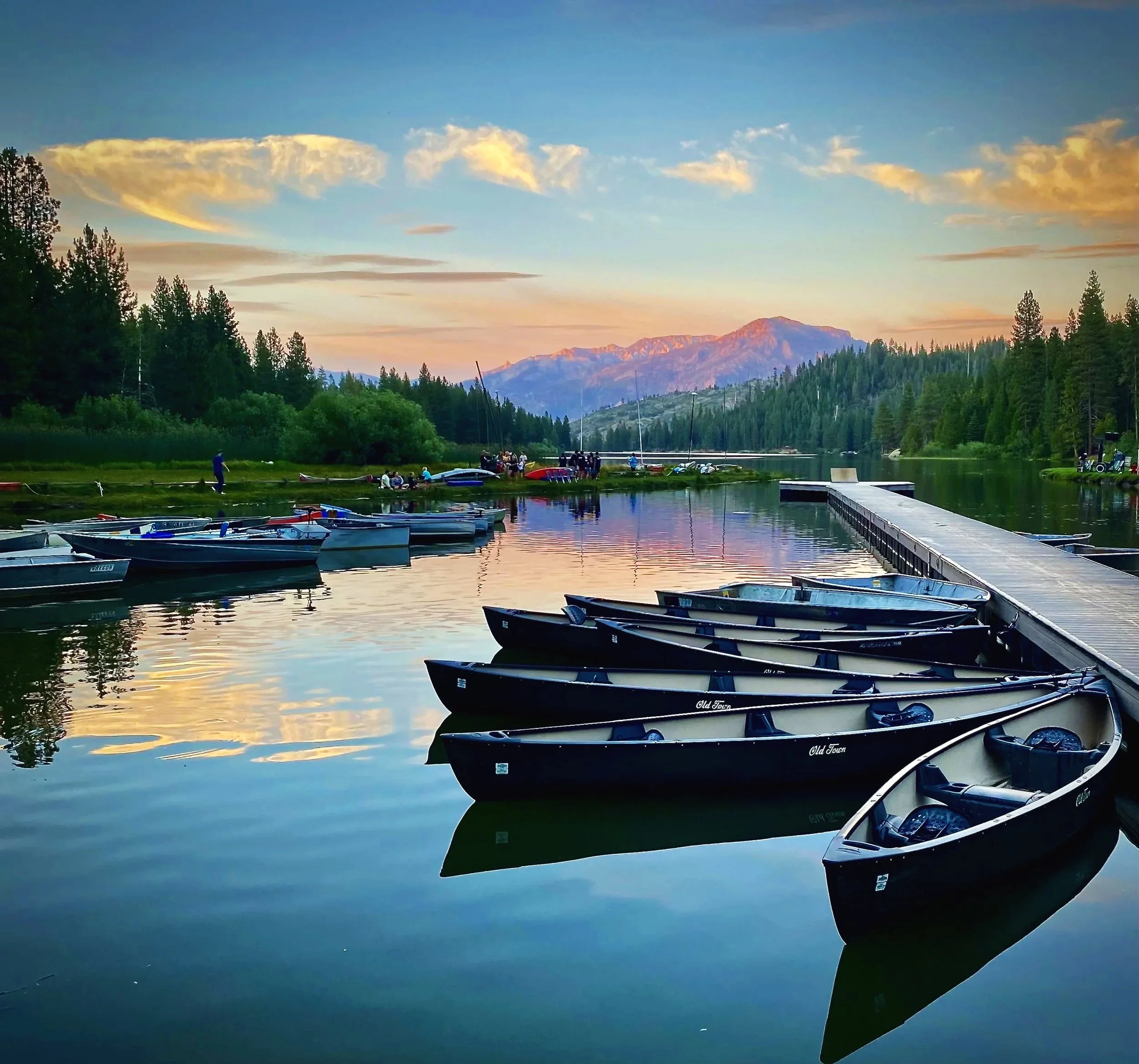 Rowboats docked along a peaceful lake at sunset with mountains and trees in the background.