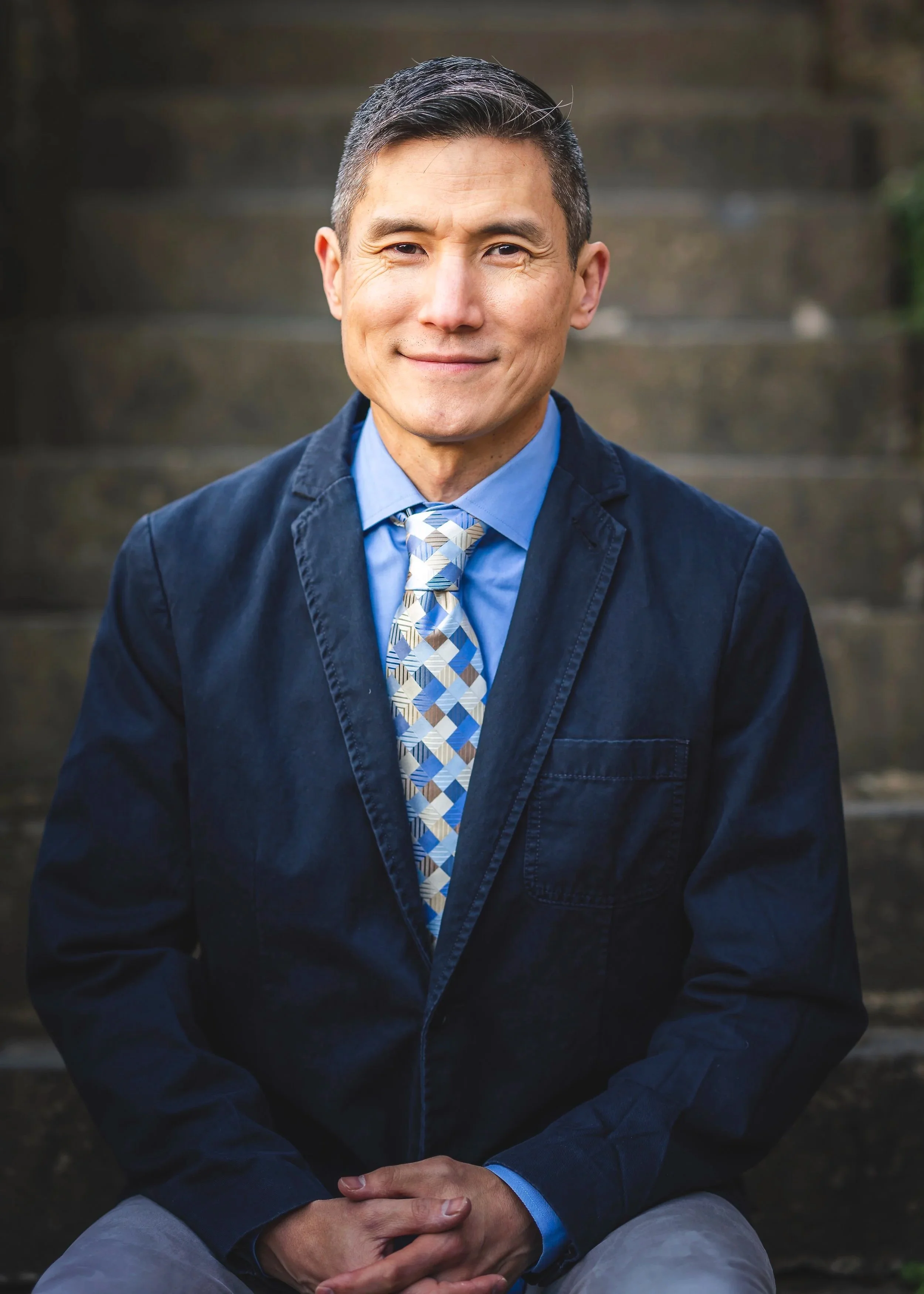 A man with short dark hair, wearing a blue shirt, a patterned tie, and a dark blazer, sitting outdoors on stone steps and smiling at the camera.