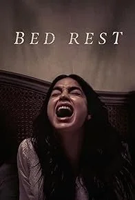 A woman with long dark hair screaming or yelling in a bedroom with the words 'BED REST' on the wall behind her.