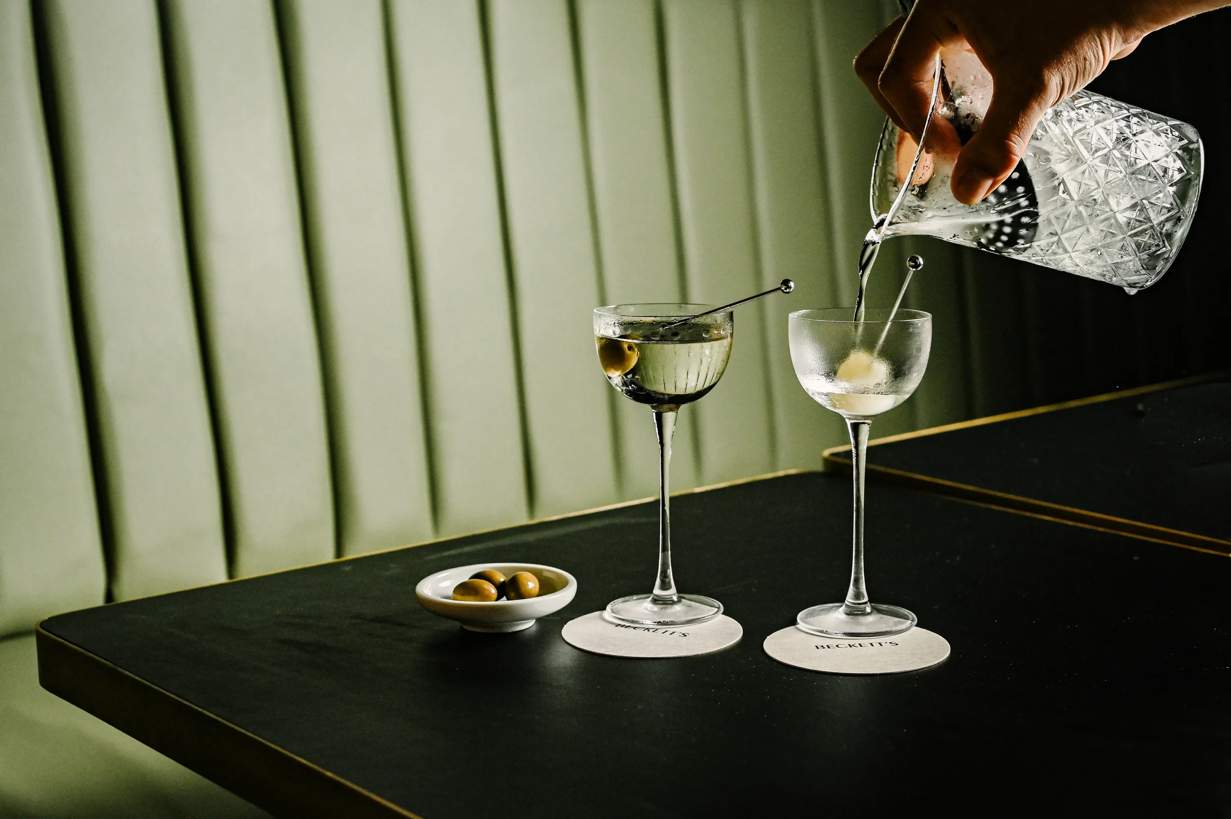 Person pouring a clear cocktail into a coupe glass with a cocktail pick, next to another coupe glass with a garnish, and a small dish of green olives on a black table with green upholstered wall in background.