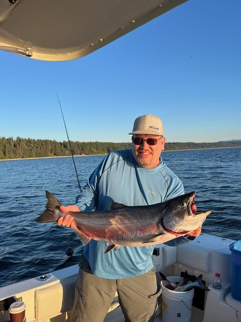 A man wearing sunglasses, a light blue long-sleeve shirt, and a white hat is smiling while holding a large fish on a boat in a body of water. The fish appears to be a large bass or similar species.