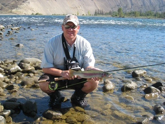 A man kneeling on rocks by a river, holding a caught fish and smiling at the camera.