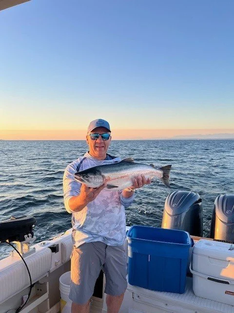 Man holding a large fish on a boat during sunset with calm water in the background.