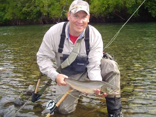 A man is kneeling in a river holding a large fish he caught, wearing fishing gear and surrounded by lush green trees.