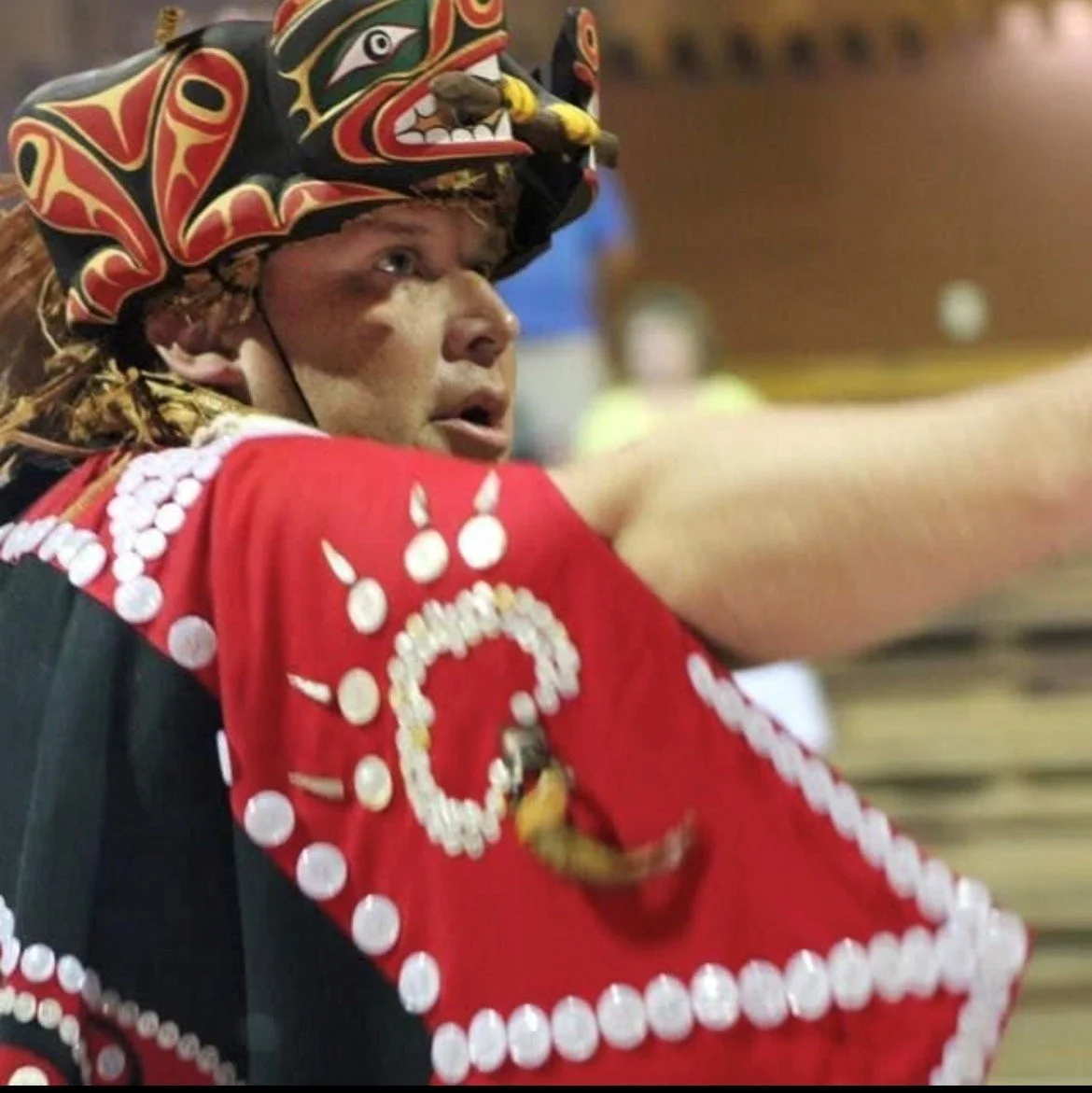 A male Indigenous athlete wearing a traditional headdress and red regalia with white decorative elements, participating in a cultural or sporting event.