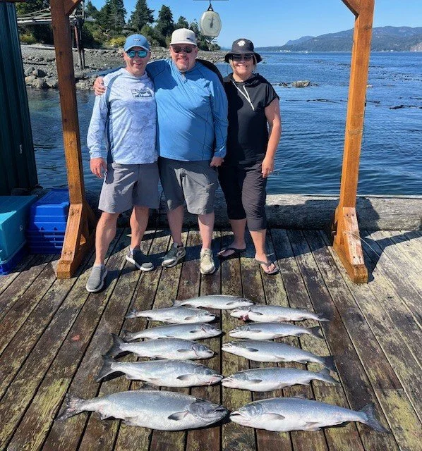 Three people standing on a wooden dock with fishing rods, smiling, with fish laid out in front of them, and the ocean and mountains in the background.