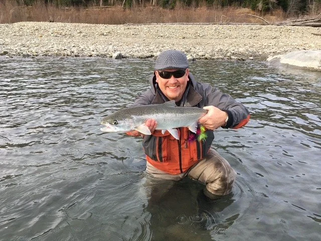 Man standing in a river holding a large fish, smiling, wearing sunglasses, a cap, and outdoor gear.