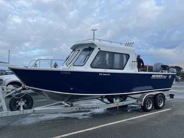 A blue and white boat on a dual-axle trailer in a parking lot, with a person standing on the boat and cloudy skies overhead.