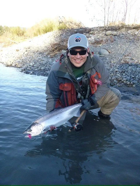 Smiling man wearing sunglasses and a cap, kneeling in a river while holding a large fish with his right hand.