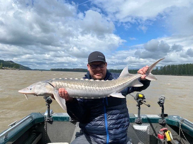 Man in a dark jacket and cap holding a large fish on a boat in a river with cloudy sky and distant trees.