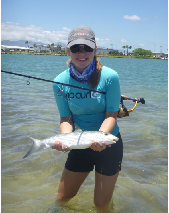 A young woman standing in shallow water holding a fish she caught, wearing a blue long-sleeve shirt, black shorts, sunglasses, and a cap, with a fishing rod on her shoulder.