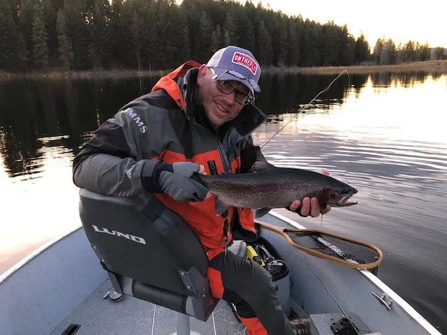 Man in outdoor fishing gear holding a fish in a boat on a lake, with a forested shoreline in the background.