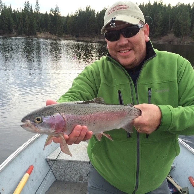 Man in a green jacket and beige cap on a boat holding a rainbow trout with a river and trees in the background.