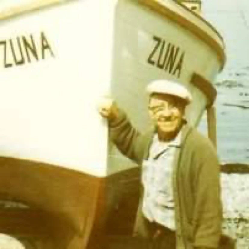 A man smiling and pointing at a boat with the word ZUNA written on it, standing near the water.