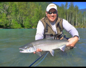Person wearing sunglasses and a cap, holding a large fish in a river with green trees in the background.