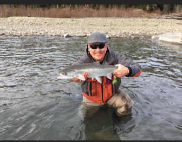 Person outdoors in a river holding a large fish, wearing sunglasses, a gray cap, and a life jacket.