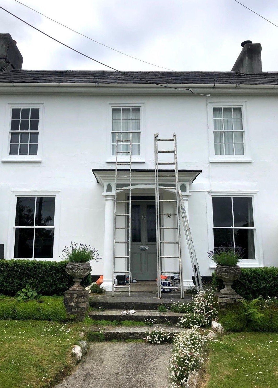 Front view of a white two-story house with a gray front door, four windows, and a small porch. Two ladders are leaning against the house, indicating ongoing maintenance or painting work. The front yard has a pathway, steps, and flower pots with plants, and is decorated with small white flowers and trimmed bushes. The sky appears cloudy.