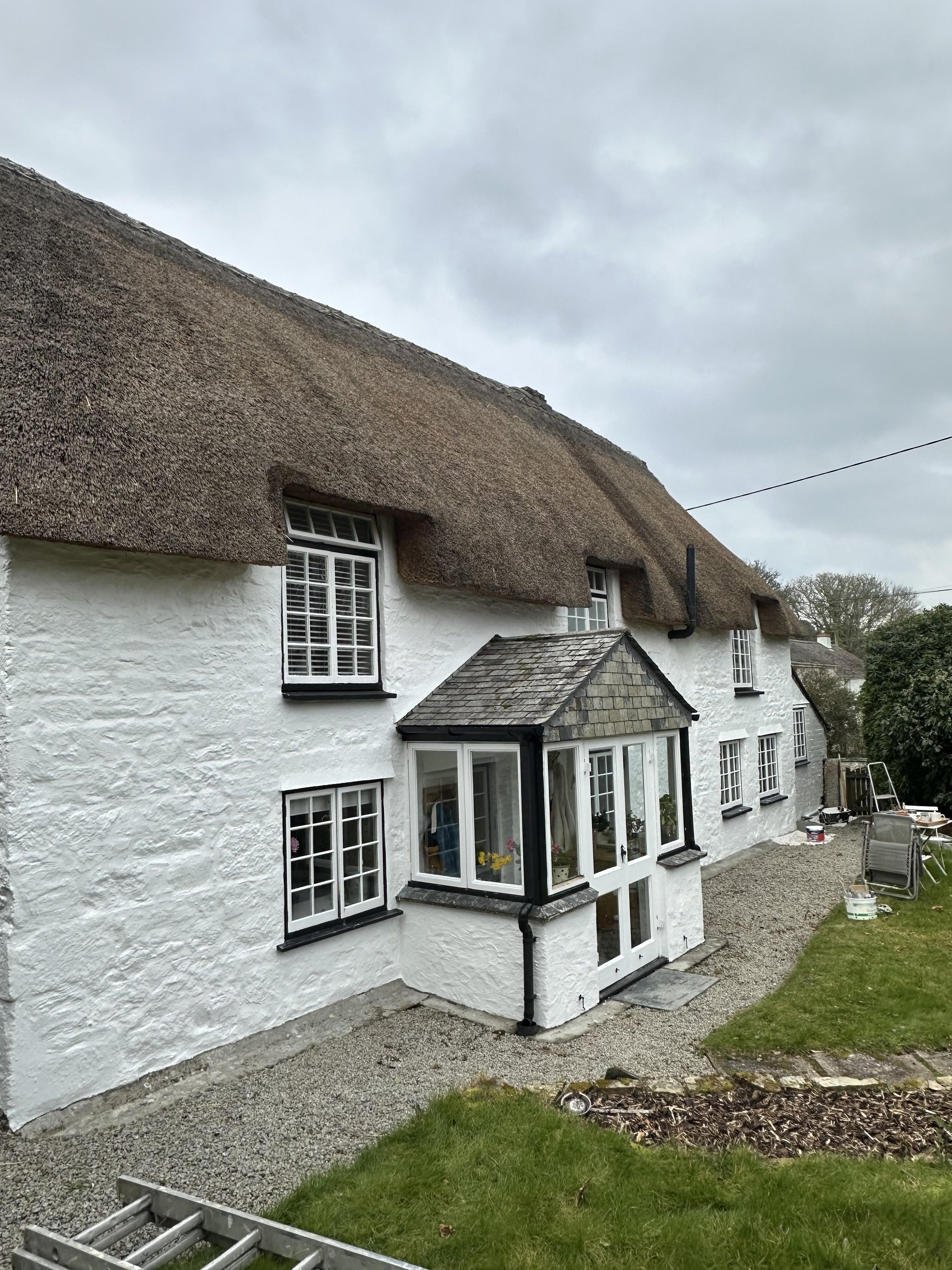 A white cottage with a thatched roof, featuring multiple windows with black trim and a glass-enclosed porch at the front. There is a gravel pathway and lawn in the foreground, with some outdoor furniture and building materials on the side.
